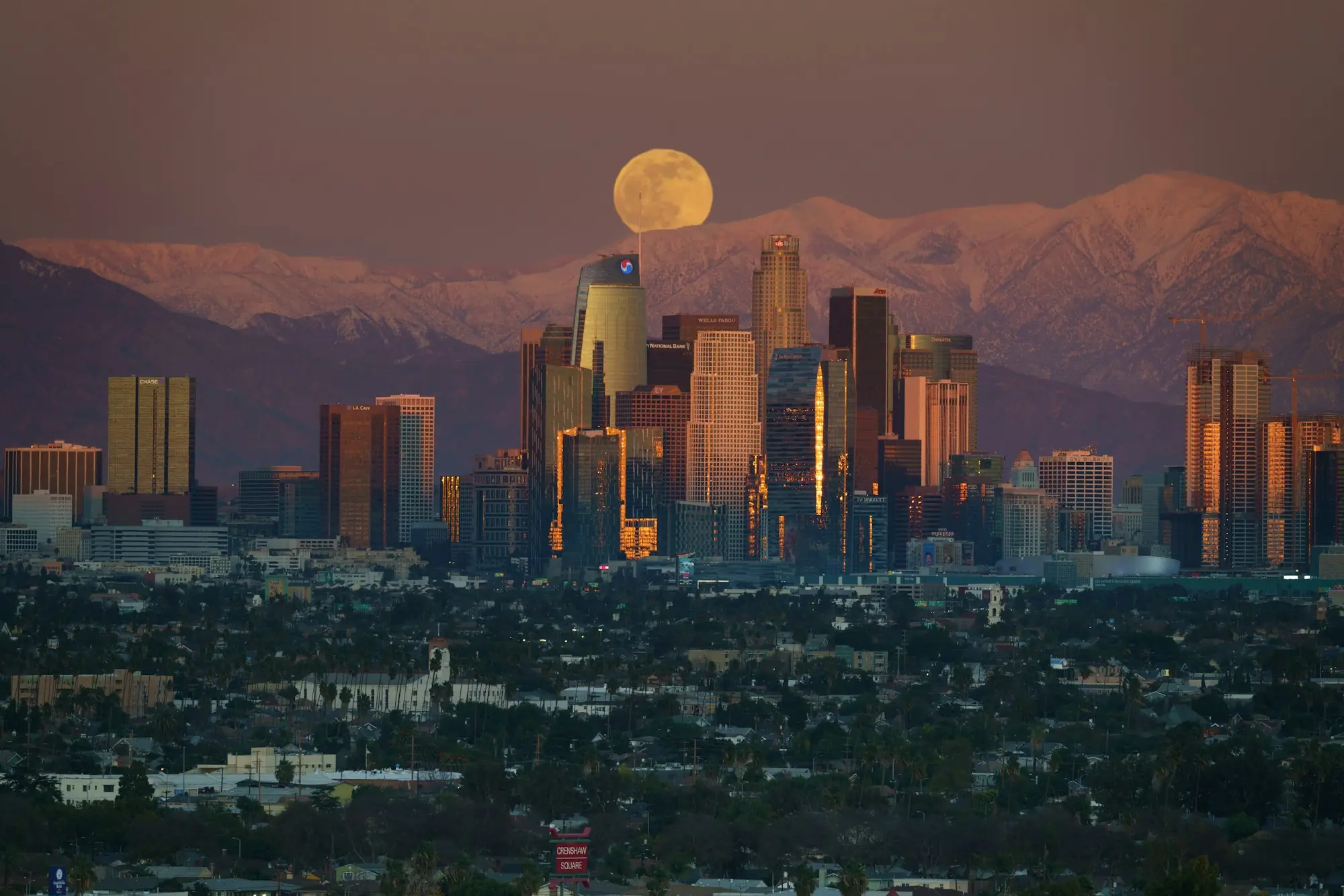 Downtown Los Angeles skyline at dusk under a full moon, with mountains behind